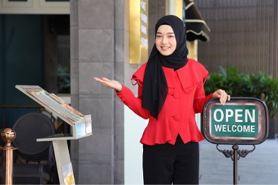 Muslim lady standing with open sign in front of restaurant.